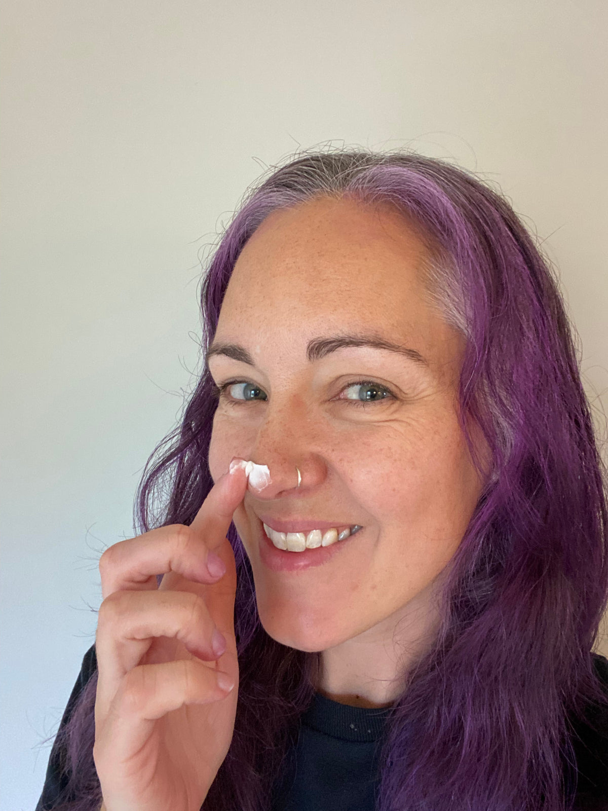 A smiling woman with purple hair applies a small white dot of moisturizer to her nose. She is wearing a silver nose ring and a black top, photographed against a plain light wall.