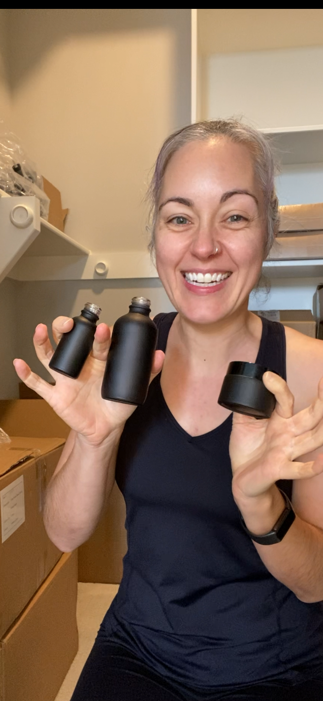 A smiling woman with gray hair pulled back, wearing a dark navy tank top, holds up three matte black cylindrical bottles in a storage room with cardboard boxes and shelves in the background.