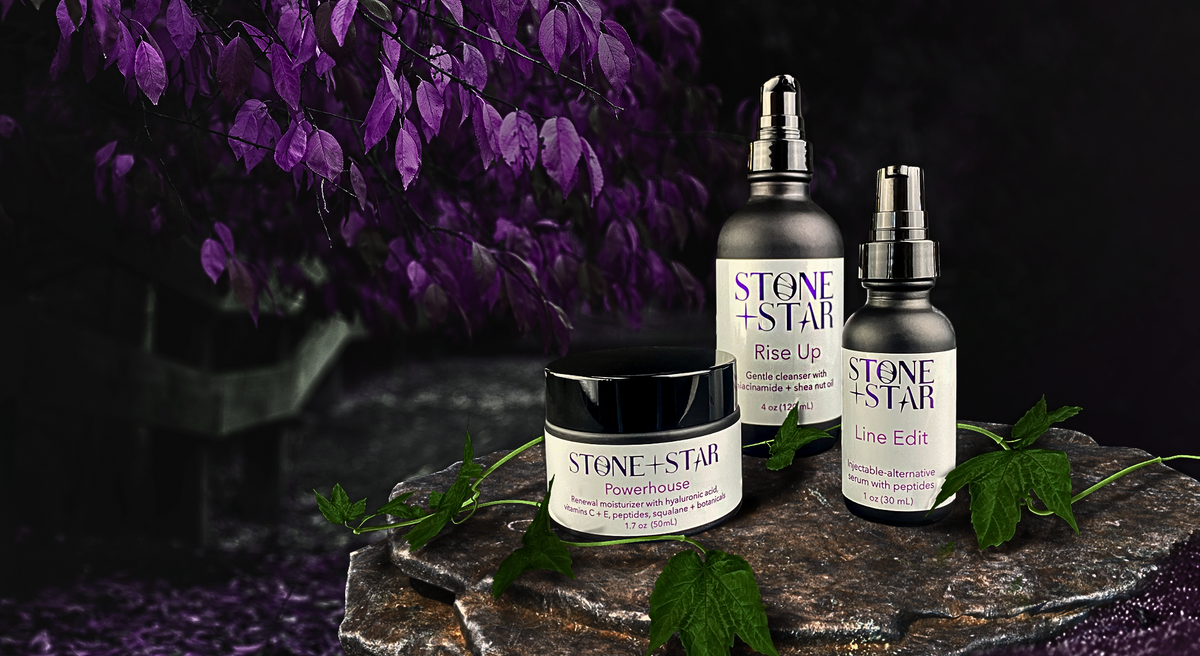 Three skincare products from Stone + Star are displayed on a dark stone pedestal with purple leaves in the background. The products include Rise Up Cleanser, Line Edit Serum, and Powerhouse Moisturizer.