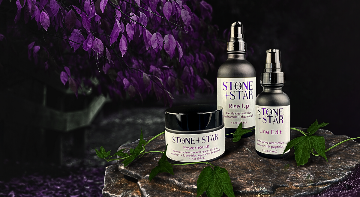 Three skincare products from Stone + Star are displayed on a dark stone pedestal with purple leaves in the background. The products include Rise Up Cleanser, Line Edit Serum, and Powerhouse Moisturizer.