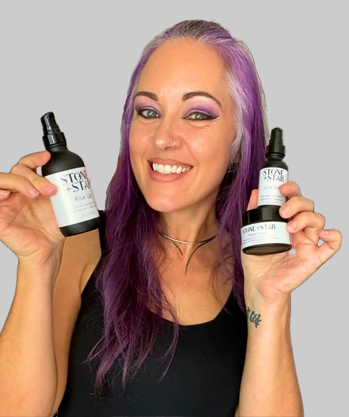 A smiling woman with grey and purple hair and a black top holds two bottles and a jar, each labeled "Stone + Star".