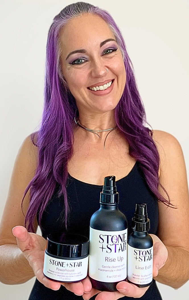 A smiling woman with long purple hair holds three black cosmetic bottles and jars against a plain light background. The products have white labels with black text reading 'STONE + STAR'.