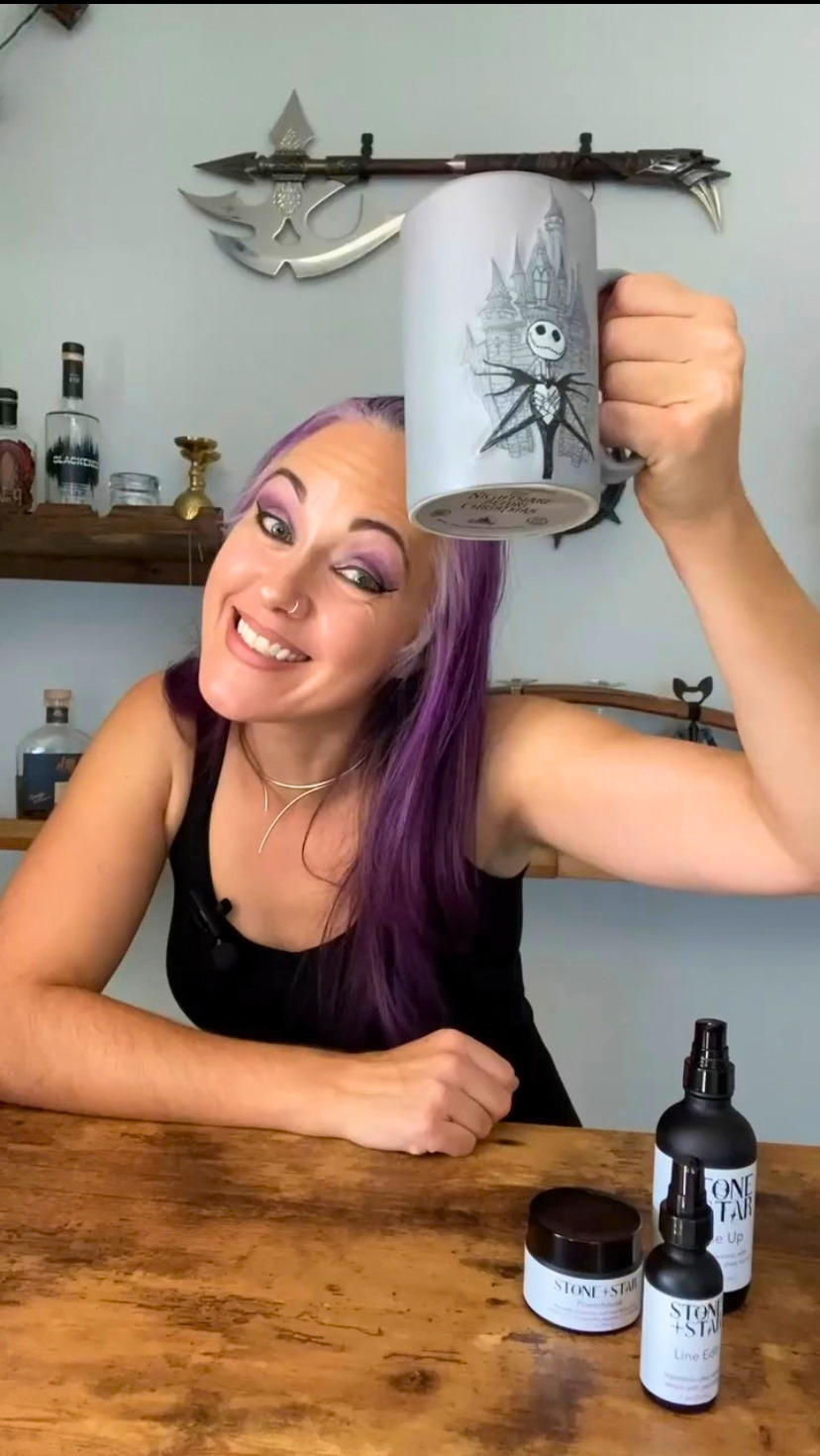 A woman with purple hair holds up a grey mug featuring a black illustration of Jack Skellington. She is sitting at a wooden table with gothic-themed bottles and jars in front of her, with a decorative axe mounted on the wall behind her.